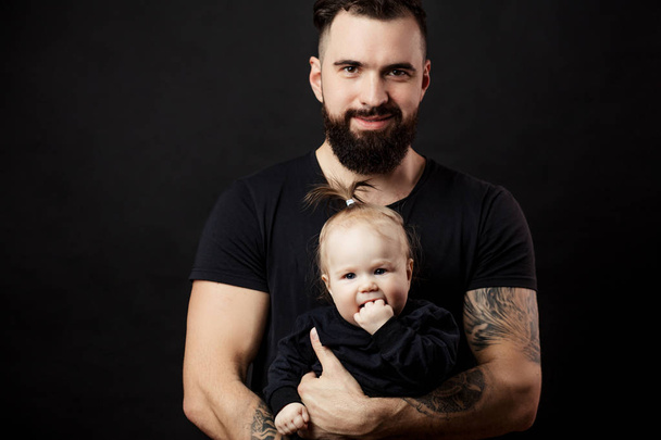 Handsome tattooed beared man holding cute little baby on black background - 写真・画像
