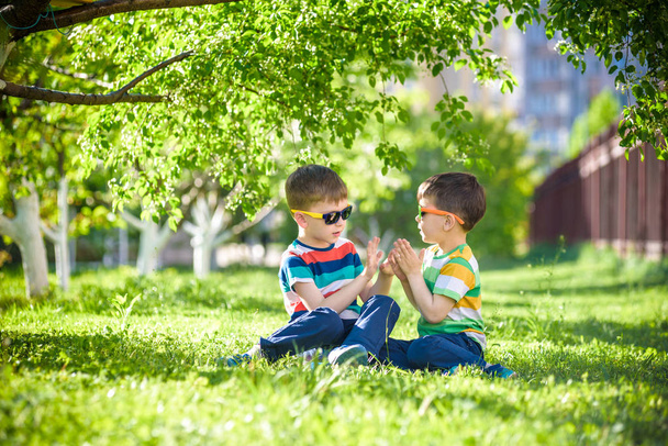 Feliz vacaciones de verano. Dos niños en un césped verde en un parque de verano. Hermanos hermanos sentados en el parque con gafas de sol sobre el fondo de los árboles florecientes. Niños feliz y sonriente concepto de amistad
. - Foto, Imagen