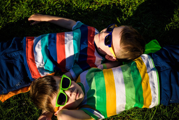 Szczęśliwe wesołe uśmiechnięte dzieci, leżące na trawie, noszące okulary przeciwsłoneczne, uśmiechnięte do aparatu fotograficznego, fotografowane z góry. Szczęśliwej koncepcji przyjaźni z dzieciństwa. - Zdjęcie, obraz