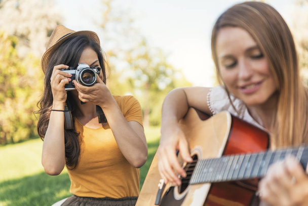 Beautiful women having fun playing guitar and photo camera in the park. Friends and summer concept. - Fotó, kép