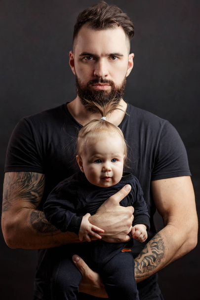 Handsome tattooed beared man holding cute little baby on black background - Foto, imagen