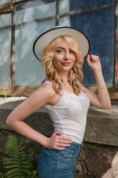 Pleased blonde woman wearing hat and stylish outfit posing near the greenhouse - 写真・画像