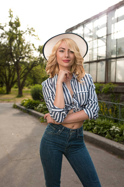 entzückendes blondes Mädchen mit Hut und stylischem Outfit posiert in der Nähe des Gewächshauses - Foto, Bild