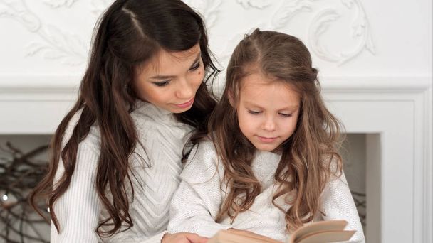 Happy mother and her beautiful daughter in white sweaters reading a book on Christmas eve - Foto, imagen