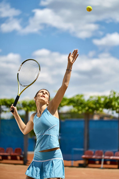 Attractive girl plays tennis on the court outdoors. She prepares to beat off a ball. Woman wears a light blue sportswear. Vertical. - Zdjęcie, obraz