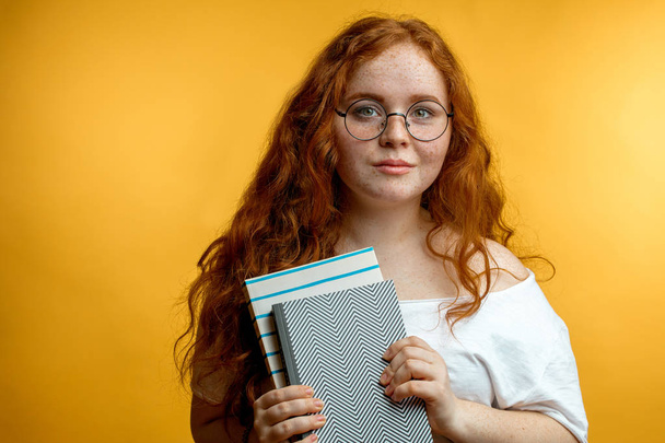 Redhead young woman holding blank books looking at the camera isolated yellow - Foto, afbeelding