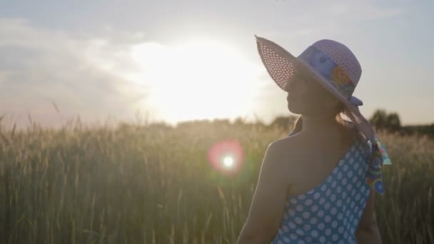 Portrait of a happy woman in a light dress and hat. The girl walks on the field with cereal plants at sunset - Footage, Video