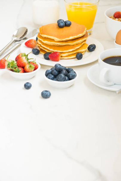 Здоровый завтрак с блинами и гранолой с черникой и клубничным и черным кофе, молоком и апельсиновым соком на белом фоне каменного стола
 - Фото, изображение