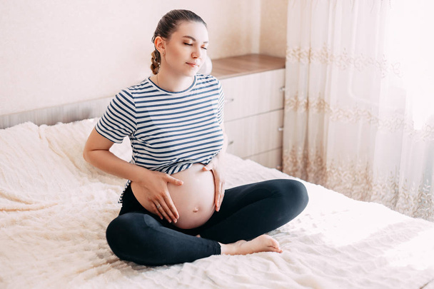 Beautiful young pregnant woman meditating on a bad in a room. - Photo, Image