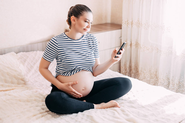 zwangerschap, moederschap, technologie, mensen en verwachting concept - close-up van zwangere vrouw met smartphone in bed thuis, met behulp van mobiele telefoon in haar slaapkamer - Foto, afbeelding
