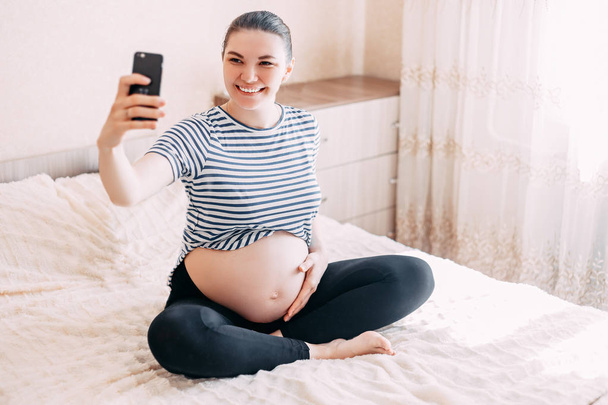 Una donna incinta che fa un selfie. Bella femmina in attesa di fare un autoritratto con il suo smartphone cercando felice
. - Foto, immagini