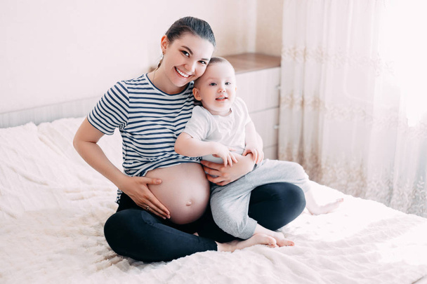 menino loiro bonito abraçando a barriga de uma mãe grávida em casa, interior. Mãe e filho brinca esperando seu segundo filho
. - Foto, Imagem