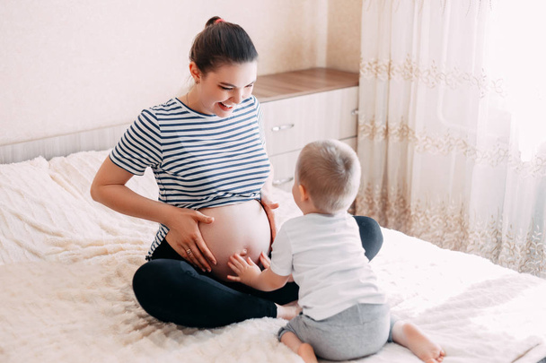 cute blonde little boy hugging a pregnant mother's belly at home, indoor. Mother and son plays in expecting their second child. - Foto, immagini