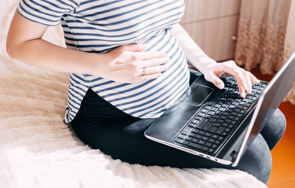 Pregnant woman working on laptop. Cropped image of pregnant businesswoman typing something on laptop while sitting at her working place at home - Fotografie, Obrázek