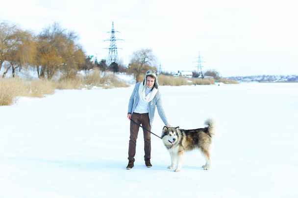 Чоловік в светрі, коричневій спідниці і рукавицях сидить на снігу і обіймає собаку хаскі
. - Фото, зображення