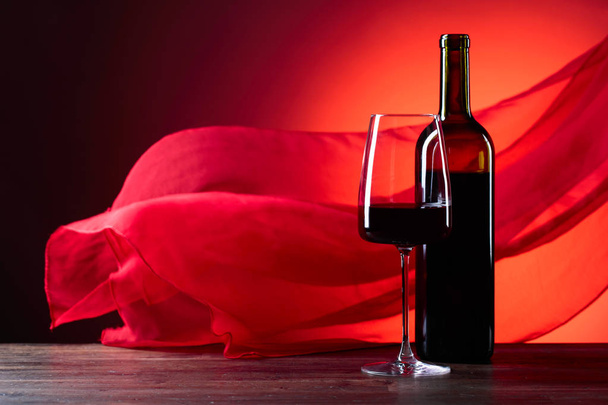 Γυαλιά και μπουκάλι κρασί rede σε κόκκινο φόντο. Κόκκινο ύφασμα σιμότητα φτερουγίζει στον άνεμο. Χώρο αντίγραφο. - Φωτογραφία, εικόνα