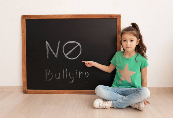 Little girl sitting near chalkboard with words "No to bullying" indoors - Photo, Image