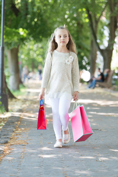 Kid shopper in fashion clothes outdoor. Child with paper bags in summer. Little princess with crown on long blond hair. Shopping on sale and purchase. Girl with shopping bags walk in park - Photo, Image