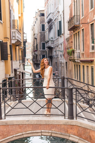 魅力的な赤毛の女性がヴェネツィアの狭い運河に橋のポーズ - 写真・画像