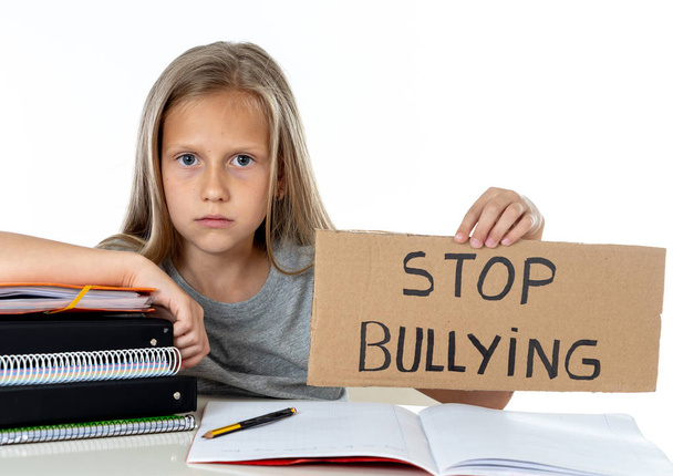young cute caucasian blonde blue eyed schoolgirl scared and stressed holding paper with text stop bullying  asking for help sitting at school desk alone.  victim children bullied abuse concept - Photo, image