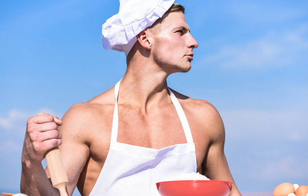 Man on confident face wears cooking hat and apron, sky on background. Cookery concept. Chef cook preparing dough for baking. Cook or chef with sexy muscular shoulders and chest holds rolling pin - 写真・画像