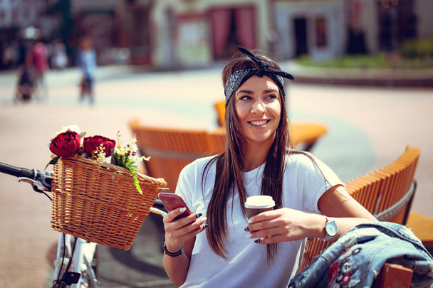 молода жінка з велосипедом з квітковим кошиком використовує смартфон і п'є каву
 - Фото, зображення