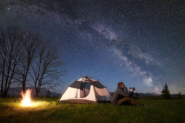 Невероятно красивые ночные лагеря в горах. Молодой человек турист с фотоаппаратом сидит в одиночестве перед туристической палаткой у горящего костра на травянистой долине под ночным голубым звездным небом с Млечным путем
 - Фото, изображение