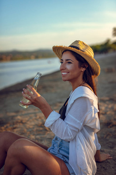 Молодая женщина в соломенной шляпе пьет пиво на пляже на закате
 - Фото, изображение