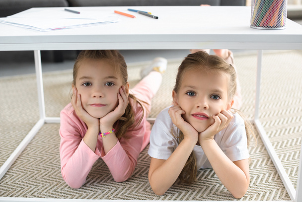portrait of little sisters looking at camera while lying under table with pencils at home - 写真・画像