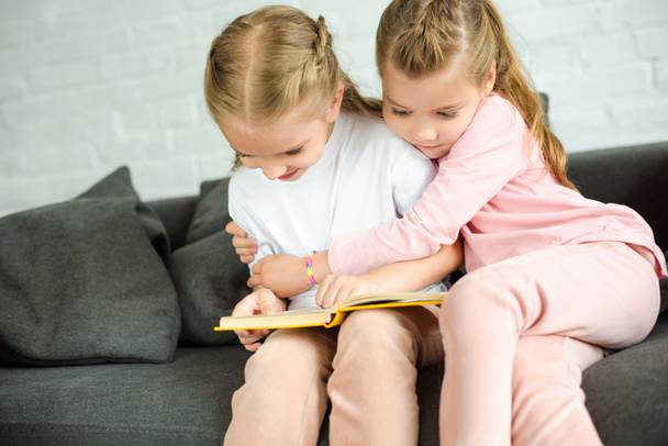 schattige kleine zusters lezen boek samen op de sofa thuis - Foto, afbeelding
