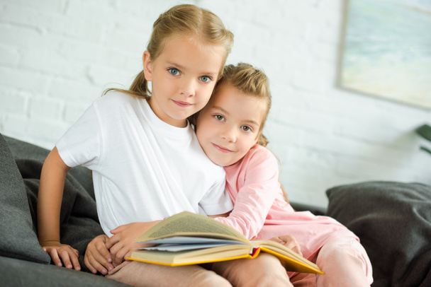 Portret van kleine kind knuffelen zus met boek op de Bank thuis - Foto, afbeelding