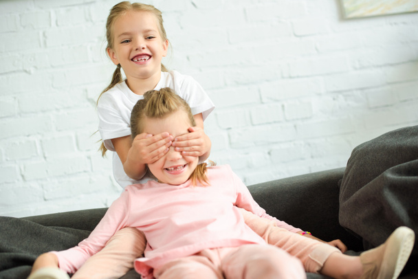 adorable little kid covering eyes to sister on sofa at home - Photo, Image