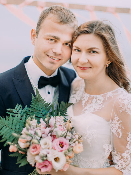 The close-up portrait of the smiling newlyweds. The bride is holding the wedding bouquet consisted of white and pink roses. - Foto, Bild