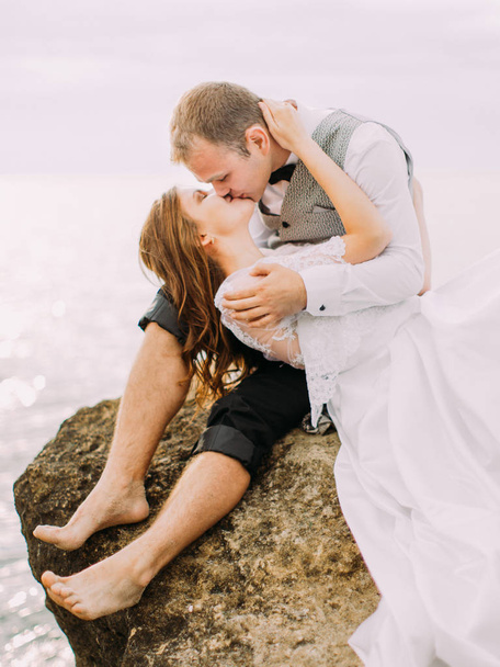 The outdoor portrait of the kissing newlyweds. The groom is sitting on the cliff while the bride is lying on his laps at the background of the sea. - Fotografie, Obrázek