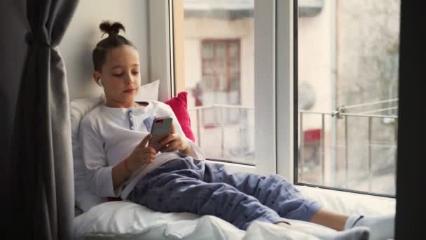 Niño sentado cerca de la ventana alféizar utiliza un teléfono inteligente y escucha música en los auriculares inalámbricos
 - Imágenes, Vídeo