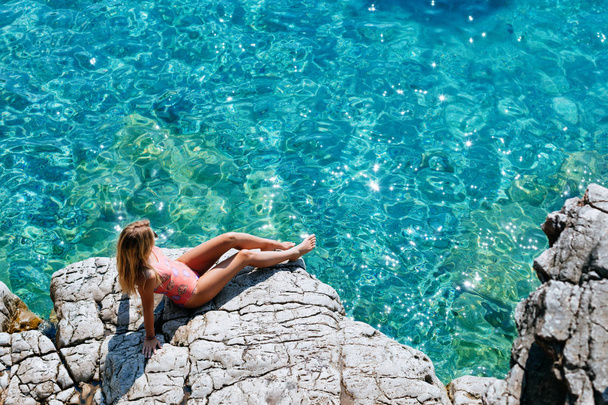 Kaunis valkoihoinen nainen istuu meren rannalla ja katselee turkoosia vettä.
. - Valokuva, kuva