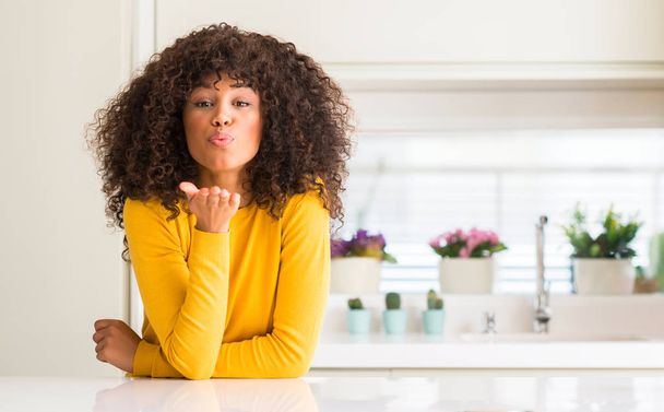 Afrikai-amerikai nő viselt sárga pulóver a konyha néz a kamerába, egy csók, kéz fúj a levegő, szép és szexi. Szeretet kifejezése. - Fotó, kép