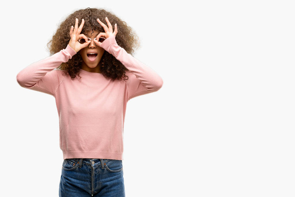 Африканская американка в розовых солнцезащитных очках делает нормальный жест, как бинокль высовывает язык, глаза смотрят сквозь пальцы. Сумасшедшее выражение
. - Фото, изображение