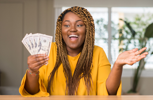 Mujer afroamericana sosteniendo dólares muy contenta y emocionada, expresión ganadora celebrando la victoria gritando con una gran sonrisa y levantando las manos
 - Foto, imagen
