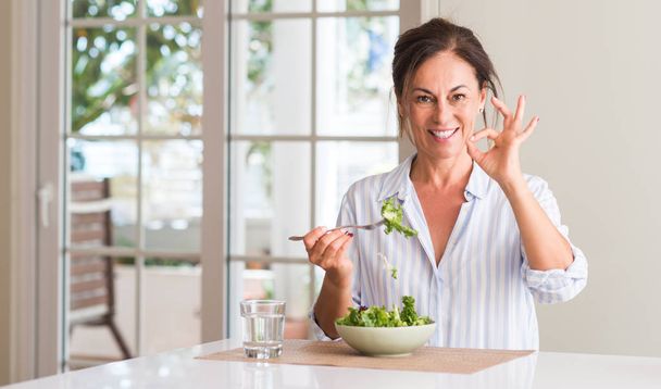 Mujer de mediana edad comiendo ensalada fresca en un tazón en casa haciendo signo ok con los dedos, excelente símbolo
 - Foto, Imagen