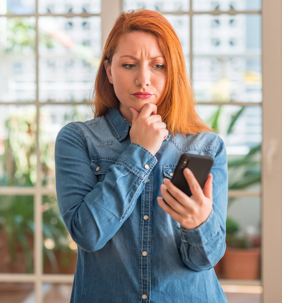 Rusovláska žena pomocí smartphone na domácí vážná tvář přemýšlet o otázku, velmi zmaten myšlenku - Fotografie, Obrázek