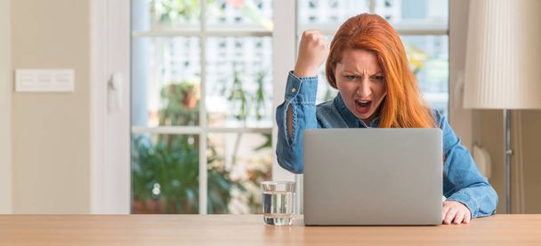 Ruda kobieta przy użyciu komputera przenośnego w domu zirytowany i sfrustrowany krzycząc z gniewem, szalony i wrzask ręce, koncepcja gniew - Zdjęcie, obraz
