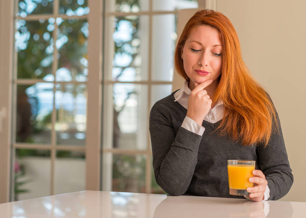 Рыжая женщина держит стакан апельсинового сока дома, серьезное лицо думает о вопросе, очень запутанная идея
 - Фото, изображение