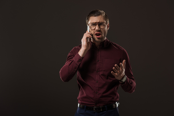 angry man in eyeglasses talking by smartphone isolated on black - Фото, изображение