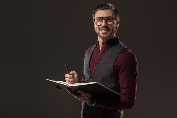 handsome buisnessman in eyeglasses taking notes and smiling at camera isolated on black - Foto, afbeelding