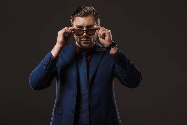 handsome stylish businessman wearing sunglasses and looking at camera isolated on black  - Fotografie, Obrázek