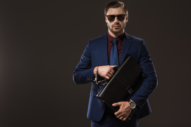 businessman in sunglasses and handcuffs holding briefcase and looking at camera isolated on black - Fotoğraf, Görsel