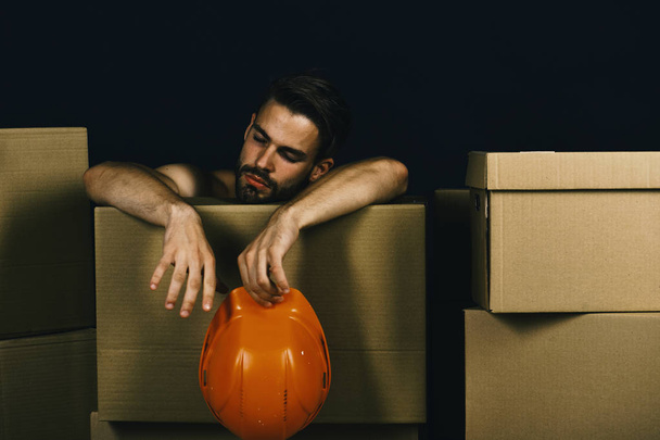 Guy with bare hands leans on boxes. Man with beard has rest on black background. Macho with tired face holds helmet. Delivery and moving - Fotoğraf, Görsel