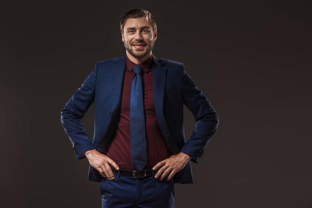 handsome businessman standing with hands on waist and smiling at camera isolated on black - Foto, imagen