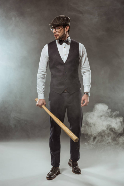 full length view of stylish man in bow tie, eyeglasses and cap holding baseball bat and looking away in smoke - Foto, immagini